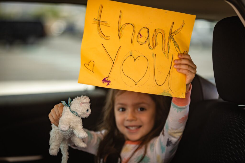A smiling child holding up a handmade 'thank you' sign and a stuffed animal while sitting in a car.